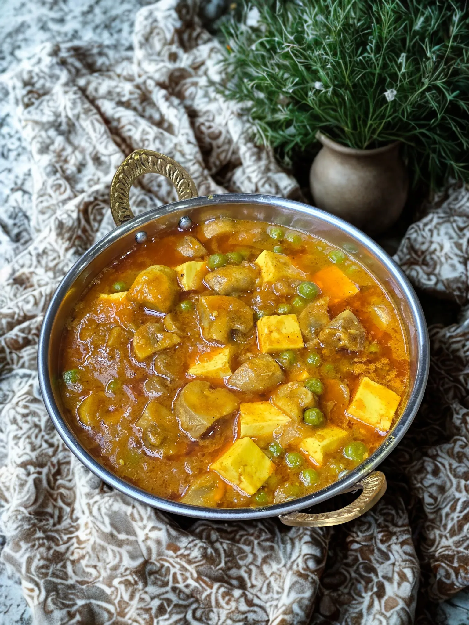 Paneer matar mushroom curry in a gold-handled kadhai on Indian block-printed fabric with a herb plant in the background